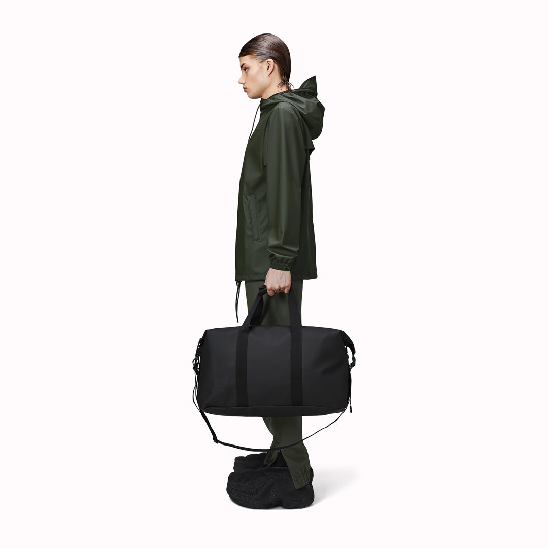 Hilo Weekend Bag by Rains as carried by model. It is a great contemporary gym or overnight bag. It features a single main compartment, carry handles, a detachable shoulder strap, and adjustable lock slider buckles on the sides. This brilliant waterproof duffel bag is cut from Rains’ signature waterproof PU coated fabric and is finished with coated zip and matte hardware.