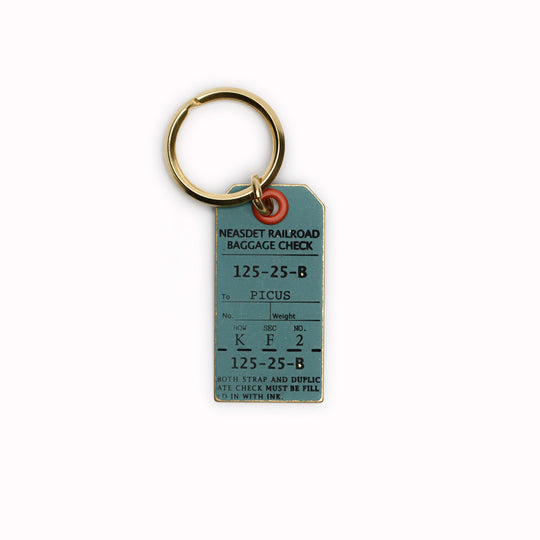 A small Japanese keychain from Picus designed to resemble an antique luggage tag and made from solid brass with a painted ash green finish and black stamped text.