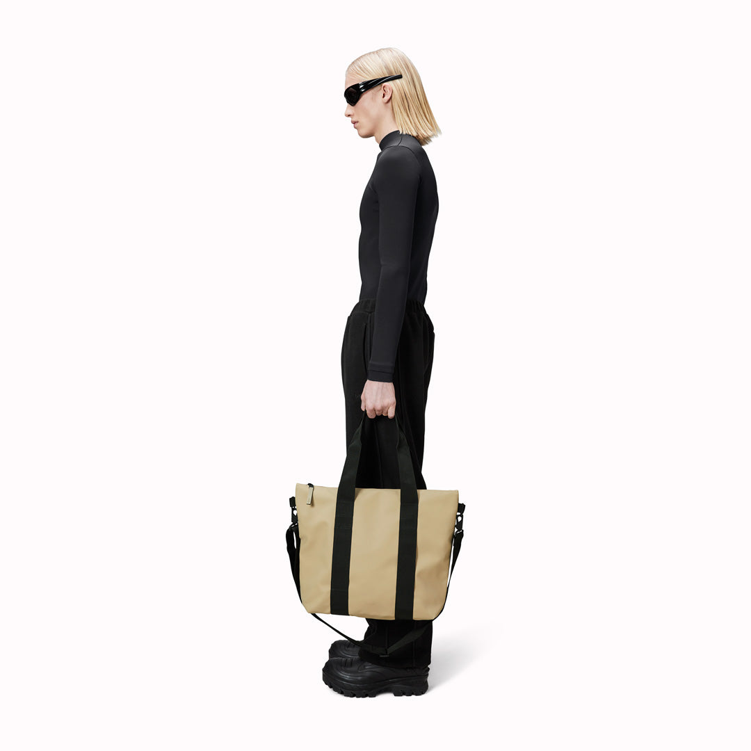 Rains' Tote Bag Mini W3 as worn by a model is a waterproof tote bag and an ideal companion for shopping trips as well as commuting to the office. Functional design elements include a full length coated zip for added water protection, which opens up to reveal a spacious main compartment. Made from Rains’ signature waterproof fabric, engineered for strength, durability and a smooth feel.