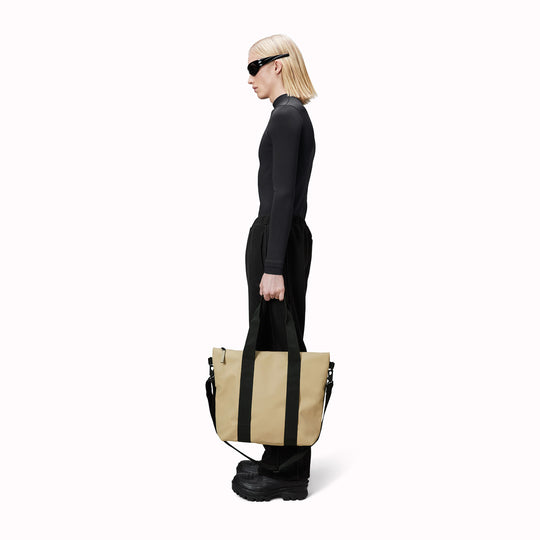 Rains' Tote Bag Mini W3 as worn by a model is a waterproof tote bag and an ideal companion for shopping trips as well as commuting to the office. Functional design elements include a full length coated zip for added water protection, which opens up to reveal a spacious main compartment. Made from Rains’ signature waterproof fabric, engineered for strength, durability and a smooth feel.