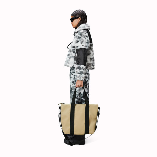 Rains' Tote Bag Mini W3 as worn by a model is a waterproof tote bag and an ideal companion for shopping trips as well as commuting to the office. Functional design elements include a full length coated zip for added water protection, which opens up to reveal a spacious main compartment. Made from Rains’ signature waterproof fabric, engineered for strength, durability and a smooth feel.