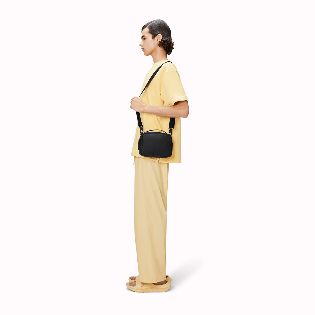 The Rains Box Bag in black is a sleek and minimalist bag crafted from Rains' signature waterproof PU fabric. It features a boxy silhouette with clean lines and a single main compartment secured by a coated zip closure.