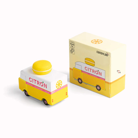 Pictured with box, the Citron Macaron Van by Candylab. It is a toy vehicle inspired by French pastry shops. It has a bright yellow color and a macaron-shaped roof rack. The van is made of solid beech wood. It is a fun and stylish addition to any child's collection.