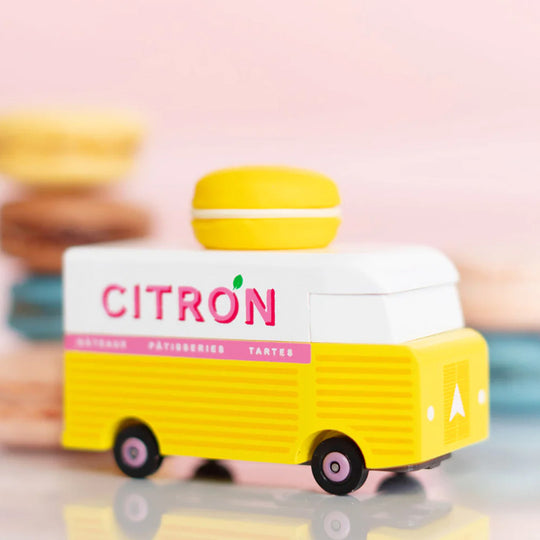 Lifestyle image on table of Citron Macaron Van by Candylab. It is a toy vehicle inspired by French pastry shops. It has a bright yellow color and a macaron-shaped roof rack. The van is made of solid beech wood. It is a fun and stylish addition to any child's collection.