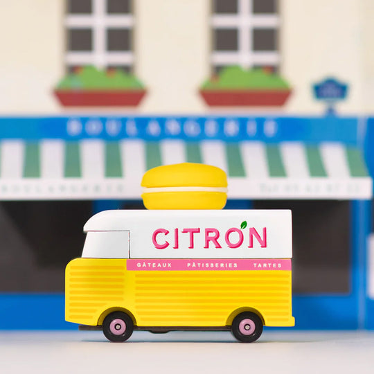 Lifestyle image, on street of the Citron Macaron Van by Candylab. It is a toy vehicle inspired by French pastry shops. It has a bright yellow color and a macaron-shaped roof rack. The van is made of solid beech wood. It is a fun and stylish addition to any child's collection.