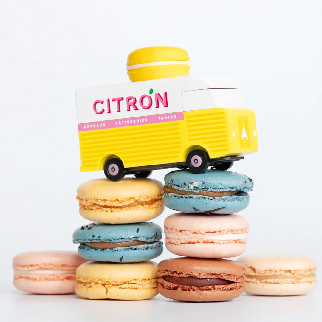 pictured on some macarons, The Citron Macaron Van by Candylab. It is a toy vehicle inspired by French pastry shops. It has a bright yellow color and a macaron-shaped roof rack. The van is made of solid beech wood. It is a fun and stylish addition to any child's collection.
