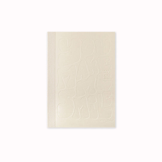 Unwrapped A6 plain paper notebook has an off white cover embossed with some ace artwork by Charlene Man featuring the words 'You Make Me Smile' in a hand-typography style. The MD paper logo is also embossed. 