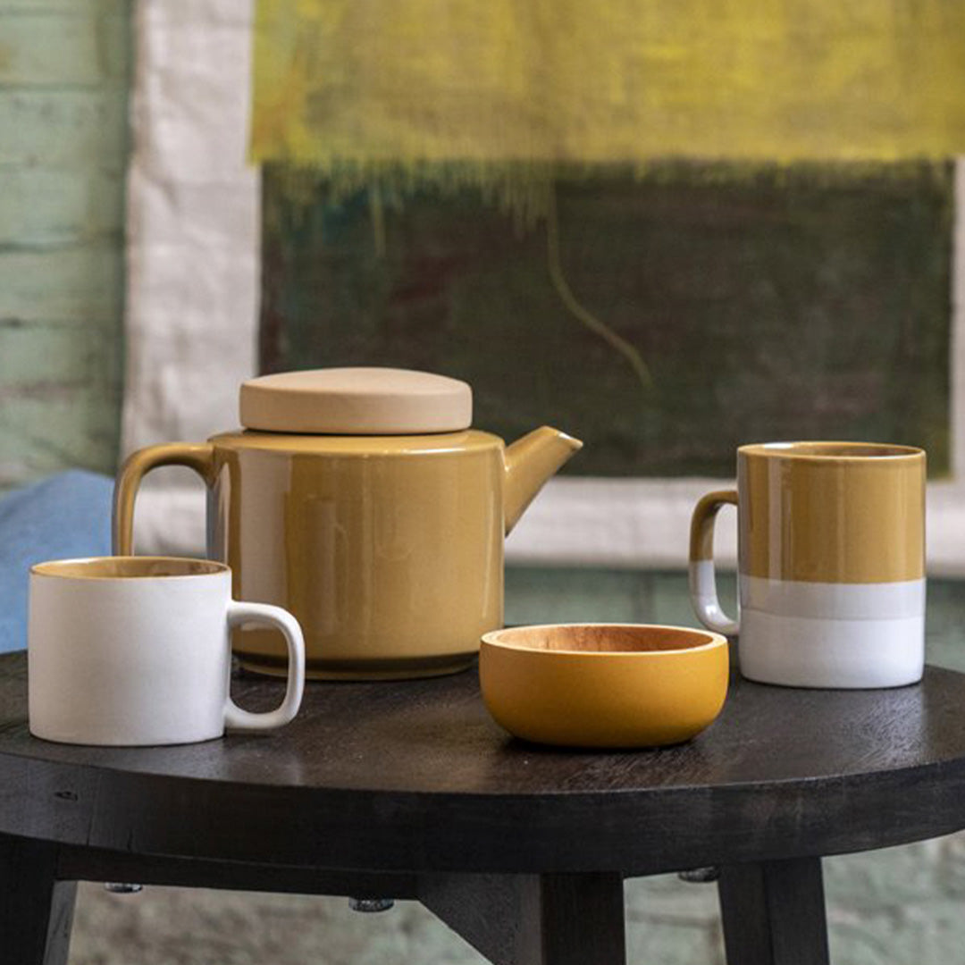 Collection on table of Large stoneware teapot from Dutch company Kinta who produce contemporary ceramics and homeware. The large teapot is mustard yellow, with a gloss glaze exterior body finish and matt glaze lid. Its design is influenced  by ceramic trends of the 1960s, but with a pleasing modern and neutral colour palette.