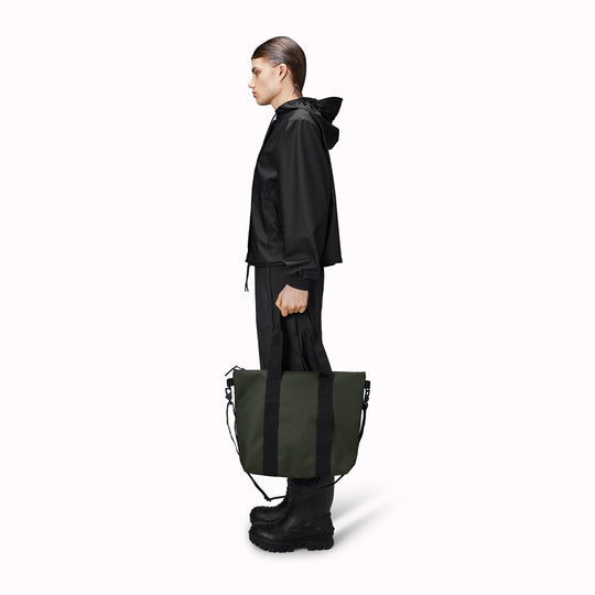 Rains' Green Tote Bag Mini W3 as worn by a model is a waterproof tote bag and an ideal companion for shopping trips as well as commuting to the office. Functional design elements include a full length coated zip for added water protection, which opens up to reveal a spacious main compartment. Made from Rains’ signature waterproof fabric, engineered for strength, durability and a smooth feel.