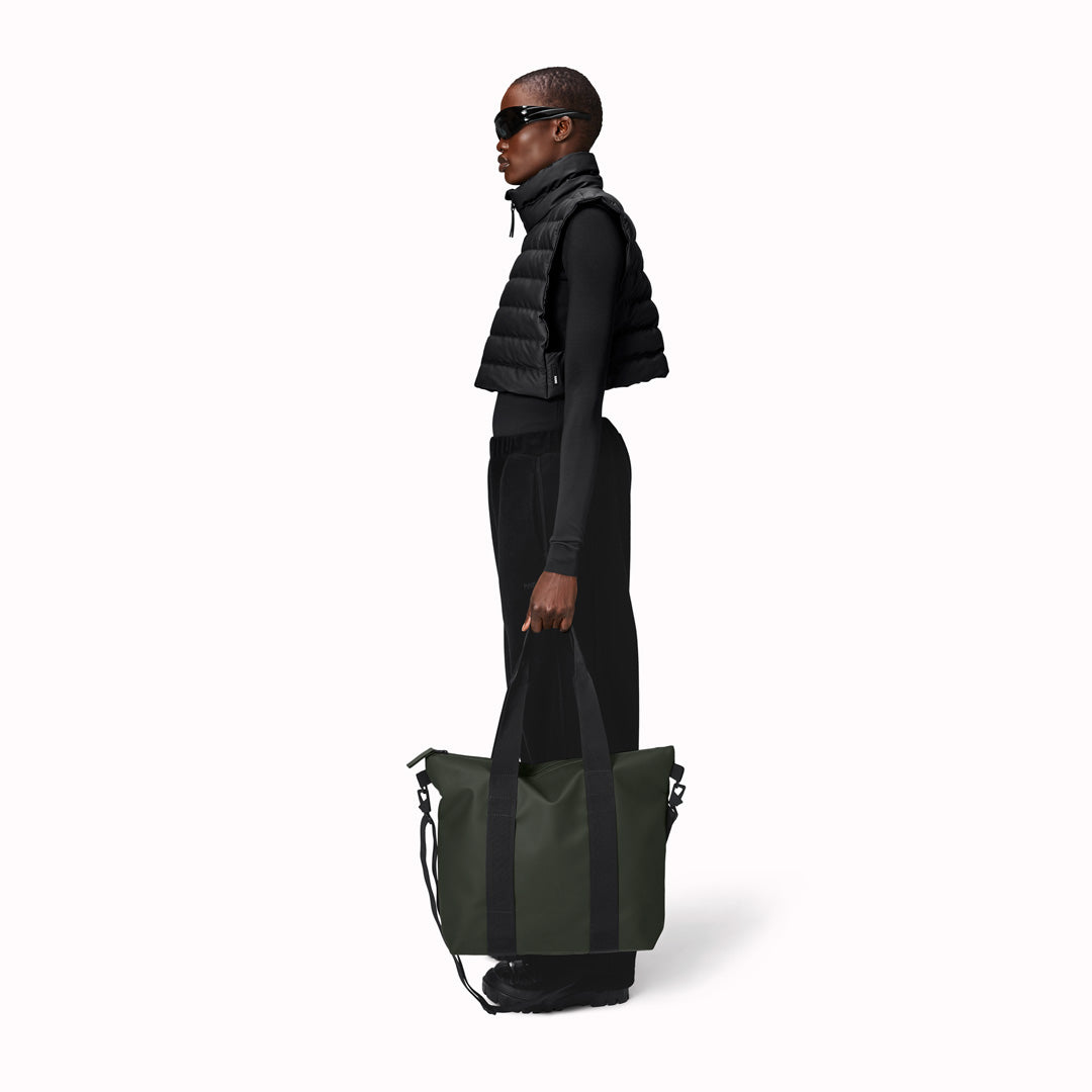 Rains' Green Tote Bag Mini W3 as worn by a model is a waterproof tote bag and an ideal companion for shopping trips as well as commuting to the office. Functional design elements include a full length coated zip for added water protection, which opens up to reveal a spacious main compartment. Made from Rains’ signature waterproof fabric, engineered for strength, durability and a smooth feel.