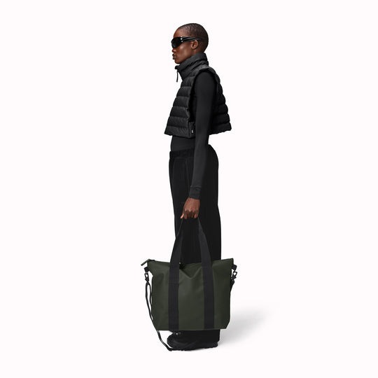 Rains' Green Tote Bag Mini W3 as worn by a model is a waterproof tote bag and an ideal companion for shopping trips as well as commuting to the office. Functional design elements include a full length coated zip for added water protection, which opens up to reveal a spacious main compartment. Made from Rains’ signature waterproof fabric, engineered for strength, durability and a smooth feel.