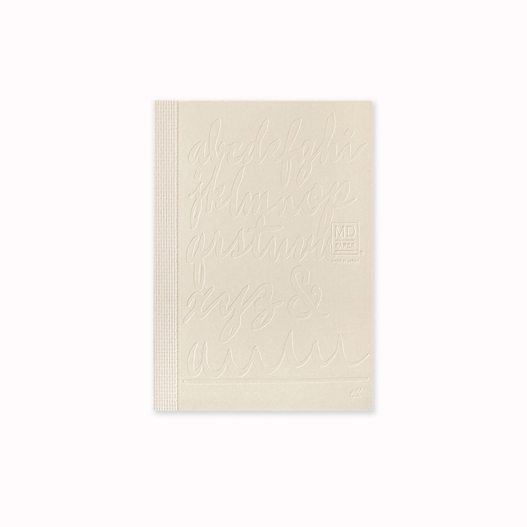 Unwrapped, This A6 plain paper notebook has an off white cover embossed with some ace artwork by Kenji Nakayama featuring a calligraphic alphabet. The MD paper logo is also embossed.  