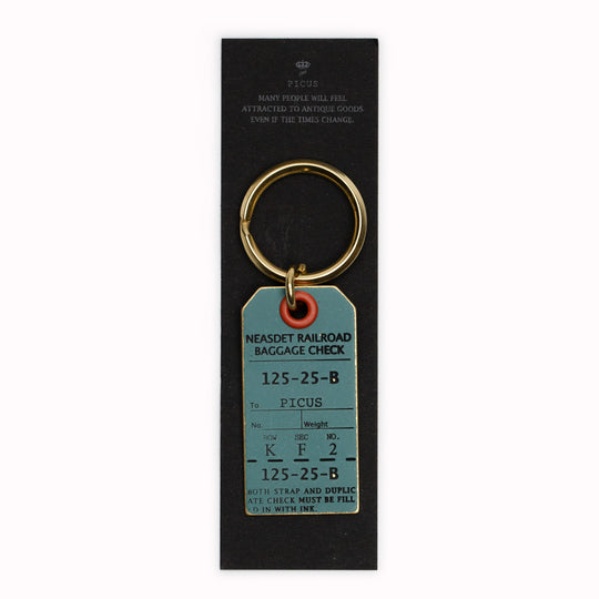 A small Japanese keychain from Picus designed to resemble an antique luggage tag and made from solid brass with a painted ash green finish and black stamped text.