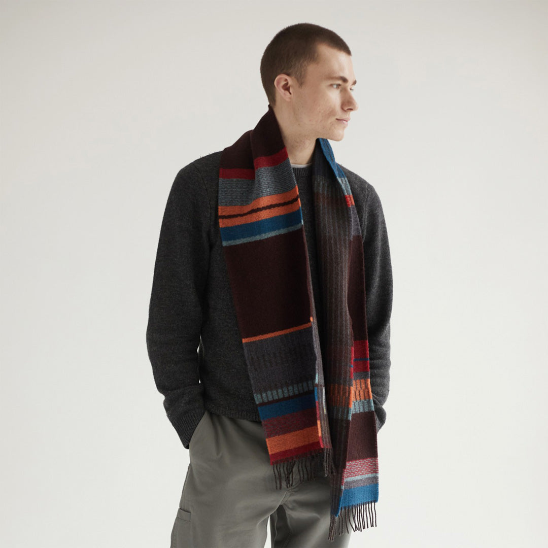 As Worn - The Strathay Cinder Lambswool scarf by Wallace and Sewell is a 100% Merino Lambswool scarf displaying a deep peat palette and berry red tones to add warmth, while the burst of orange adds a lively touch.