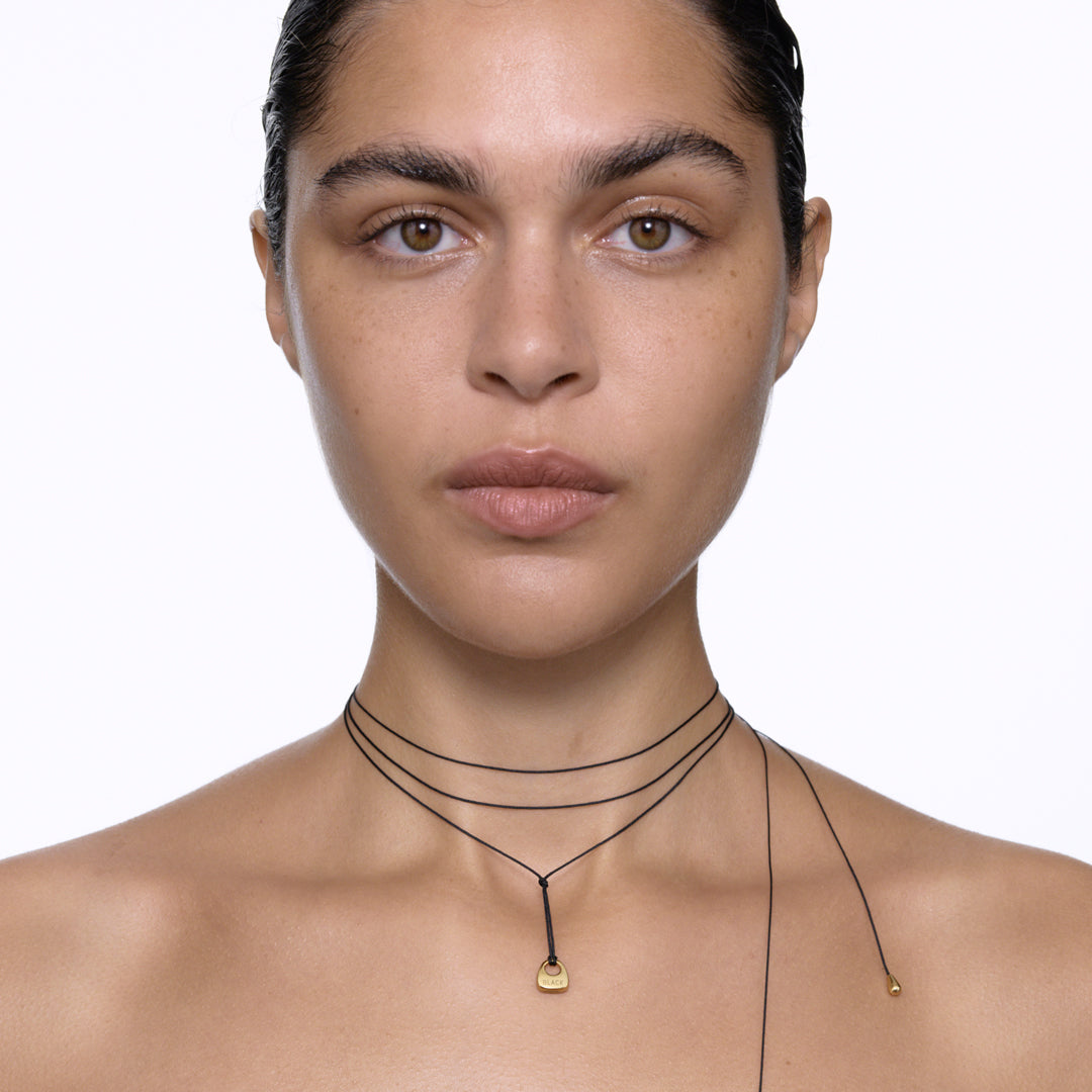 Maria Black's Tote Necklace, crafted in recycled sterling silver or 18ct gold plating features a soft trapeze-shaped pendant 'Tote' engraved with “BLACK,” it hangs from a 170cm adjustable cord made from recycled polyester.