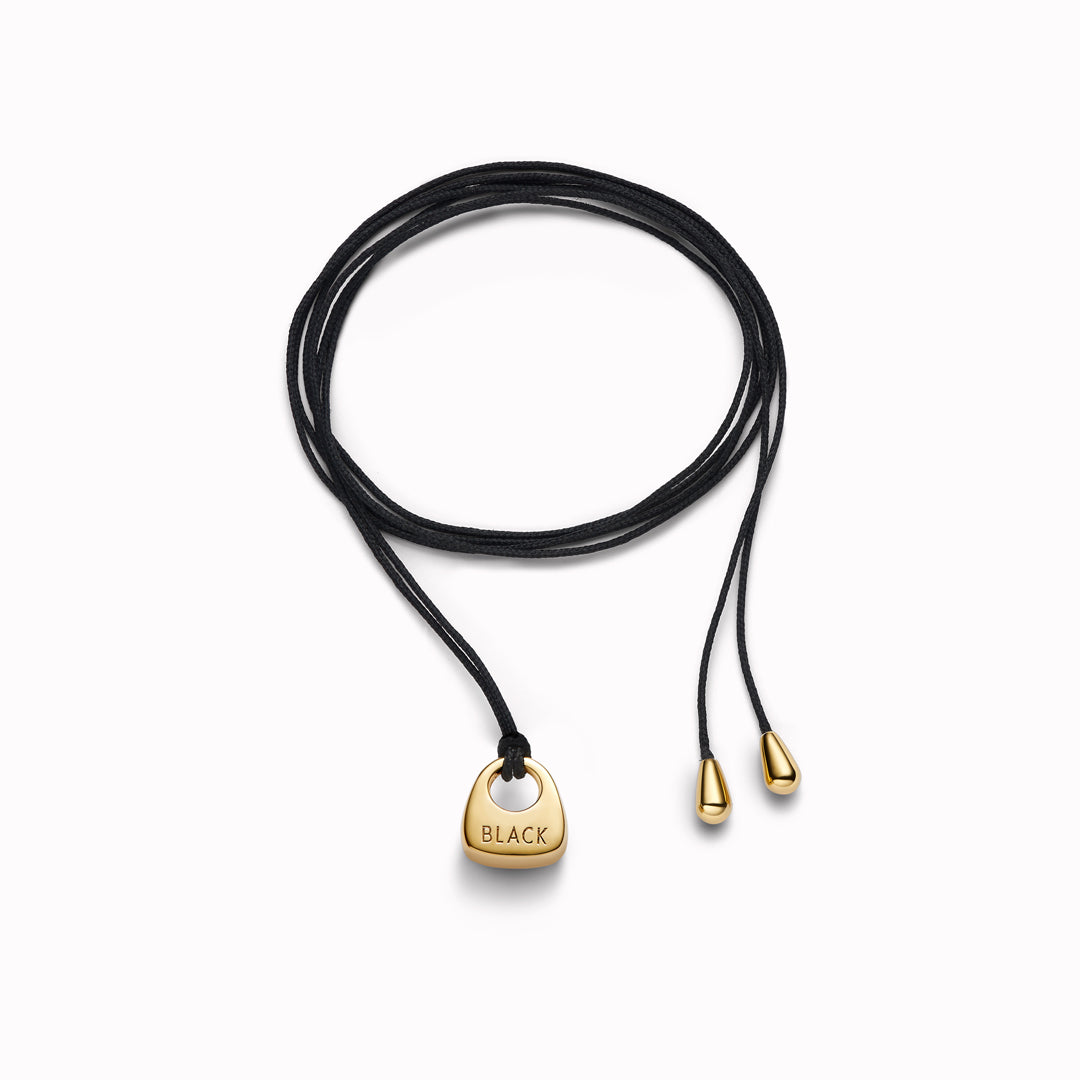 Maria Black's Tote Necklace, crafted in recycled sterling silver or 18ct gold plating features a soft trapeze-shaped pendant 'Tote' engraved with “BLACK,” it hangs from a 170cm adjustable cord made from recycled polyester.