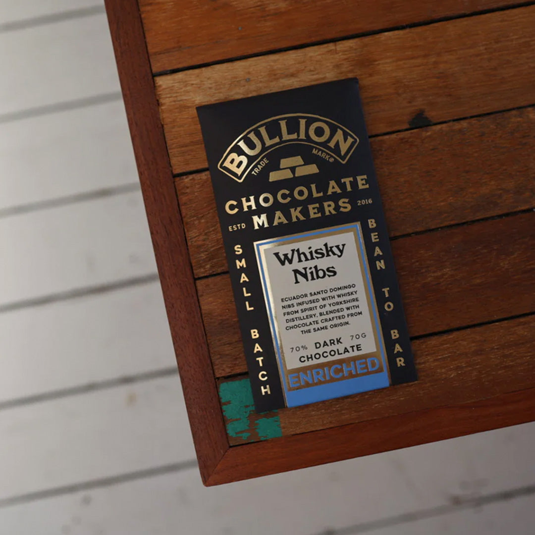 The Whisky Nib Bar from Bullion contains Ecuadorean Santo Domingo nibs infused with whisky from Spirit of Yorkshire Distillery, blended with chocolate crafted from the same origin. Shown on table.