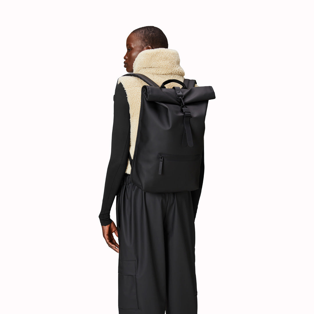 As Worn by model. Black Rolltop Rucksack. Made from Rains’ signature waterproof fabric, this functional backpack has a roll-top closure with an adjustable strap featuring a loop for a bike lock or similar. It has a large main compartment, and an easy-access front pocket and side-access laptop compartment both with a water-repellent zipper closure. 