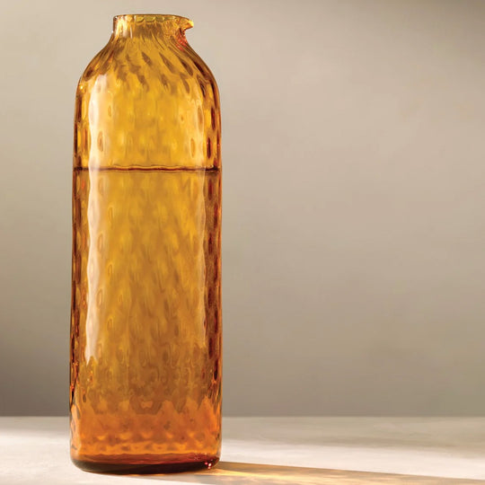 The Dapple Carafe is a lattice textured bottle shaped carafe made from coloured amber yellow mouth-blown glass. The Dapple Carafe features a beautiful undulating textured surface that is inspired by dappled sunlight on water.