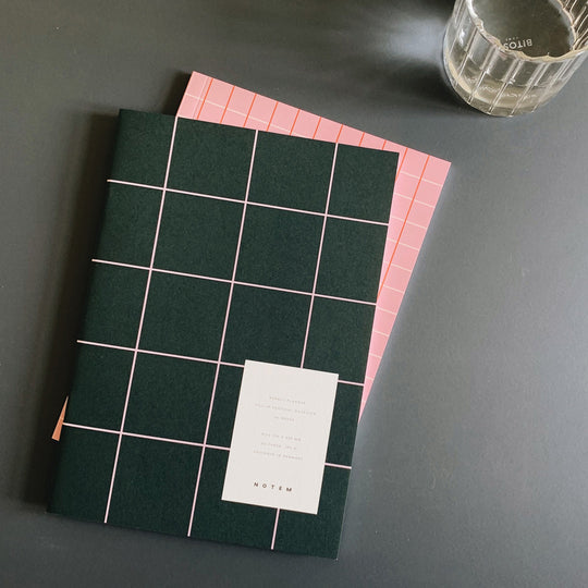 The planner contains 23 undated weekly spreads and has a horizontal layout with space for notes and must-dos of the week. It is made of high-quality uncoated paper and has a soft cover in light grey or dark green with a grid pattern.