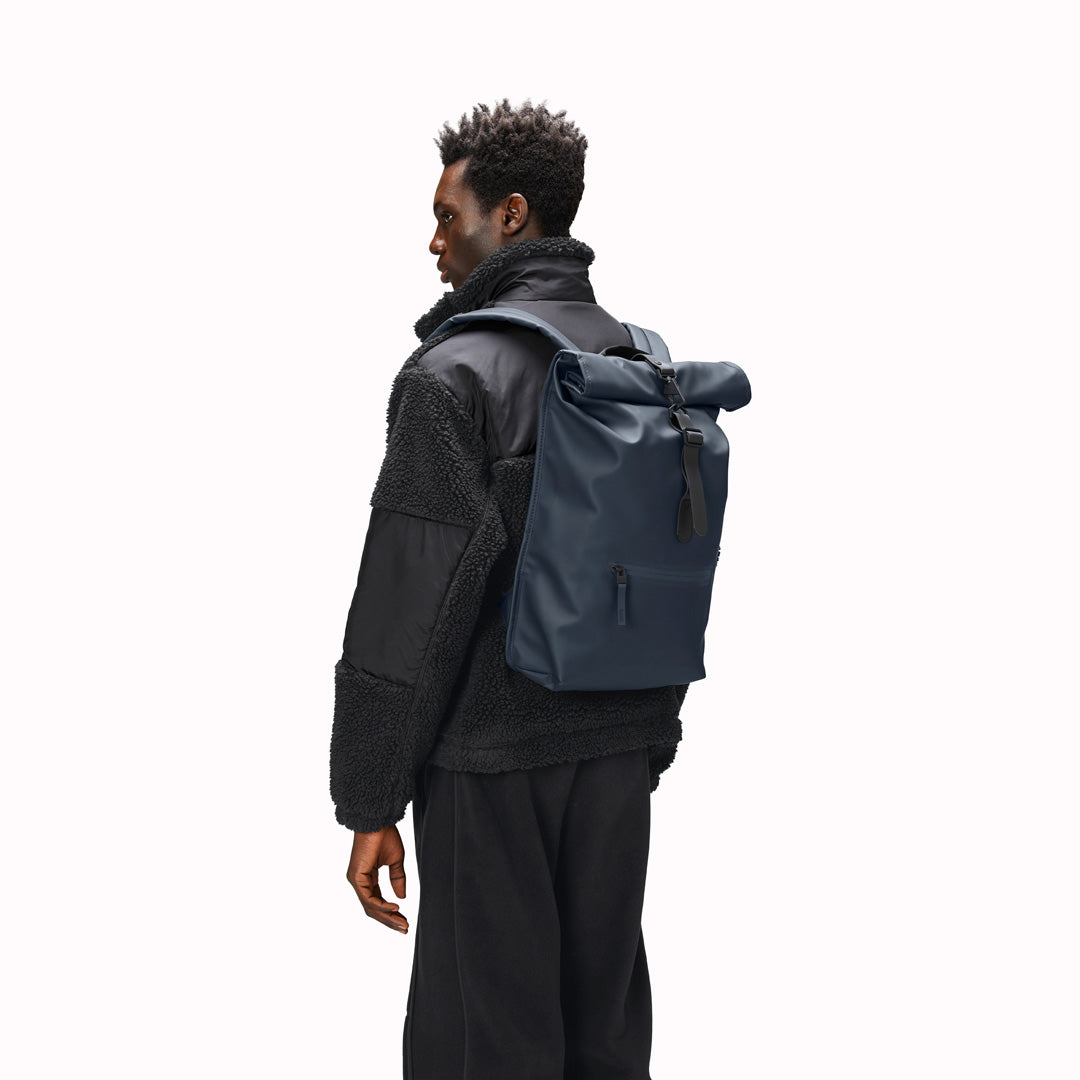 As worn by model. View of Navy Rolltop Rucksack. Made from Rains’ signature waterproof fabric, this functional backpack has a roll-top closure with an adjustable strap featuring a loop for a bike lock or similar. It has a large main compartment, and an easy-access front pocket and side-access laptop compartment both with a water-repellent zipper closure.