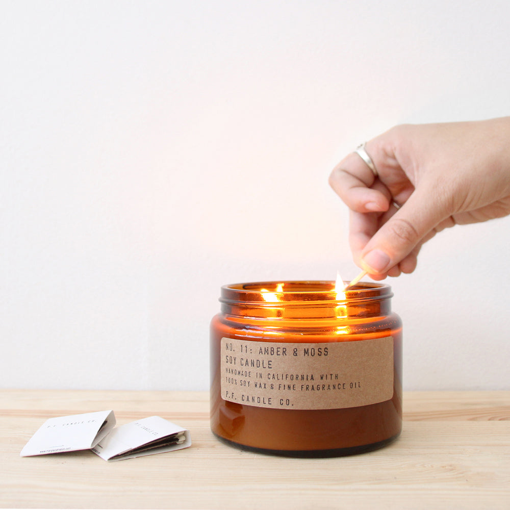 PF Candle Amber and Moss, Small jar being lit on table. These classic candles from P.F. Candle Co are hand poured into apothecary inspired amber jars with signature kraft label and brass lid. A warm, comforting glow and divine scent.