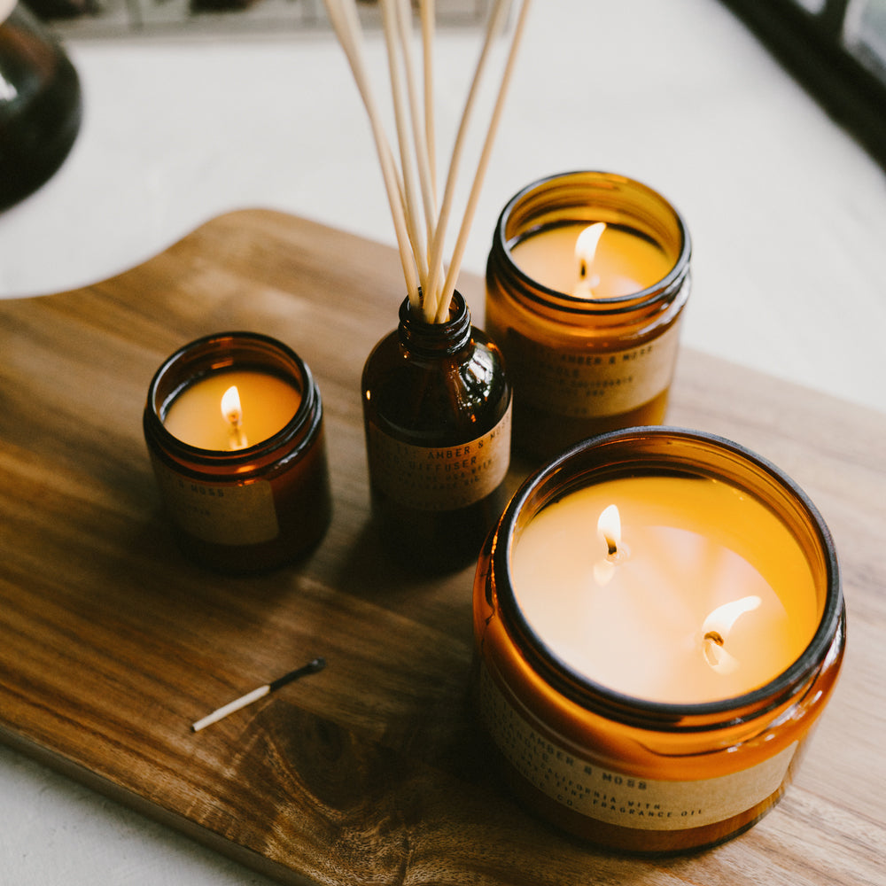 PF Candle Amber and Moss collection on table image. These classic candles from P.F. Candle Co are hand poured into apothecary inspired amber jars with signature kraft label and brass lid. A warm, comforting glow and divine scent.