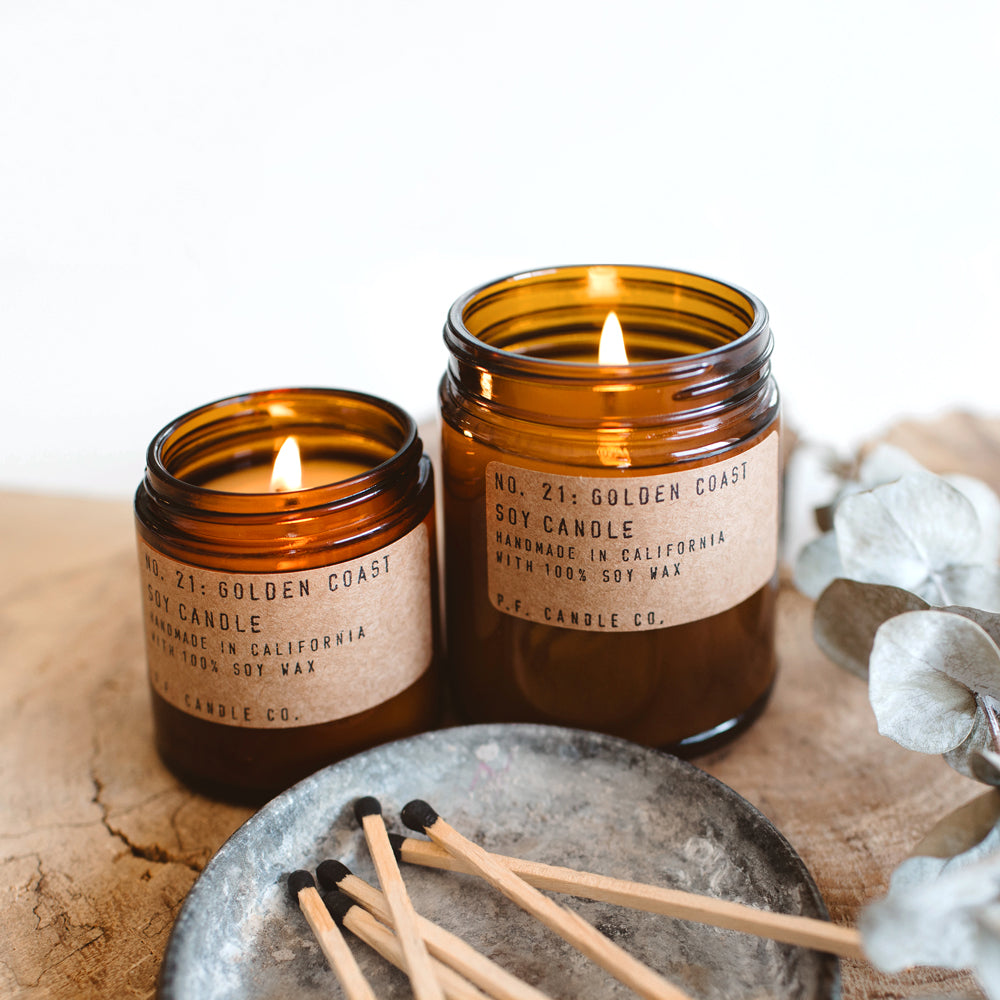 PF Candle Golden Coast, Small jar being lit on table. These classic candles from P.F. Candle Co are hand poured into apothecary inspired amber jars with signature kraft label and brass lid. A warm, comforting glow and divine scent.