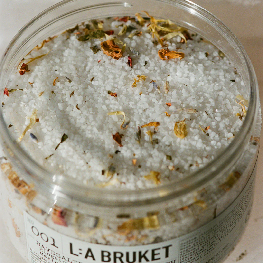 Marigold, Orange and Geranium Sea Salt Bath Detail | 001 | La Bruket with lid off, showing petals in salt bath. A sea salt bath enriched with marigold, orange and geranium. The base of sea salt cleanses and stimulates the skin. Contains essential oil of marigold, orange and geranium which has calming, antibacterial and repairing properties to effectively counteract skin irritations. Organic and/ or natural ingredients. Natural Swedish self-care from L:A Bruket.