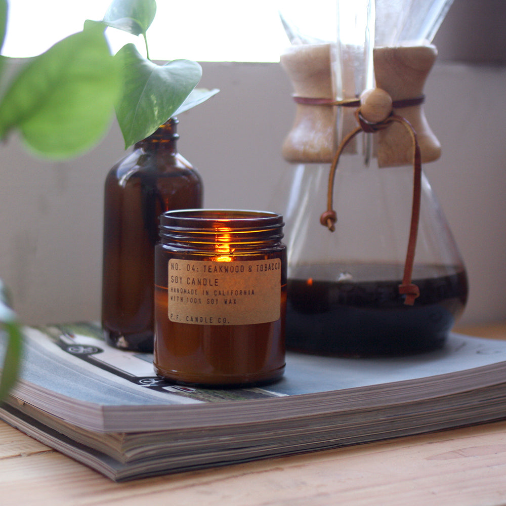 PF Candle Teakwood and Tobacco, Small jar being lit on table. These classic candles from P.F. Candle Co are hand poured into apothecary inspired amber jars with signature kraft label and brass lid. A warm, comforting glow and divine scent.