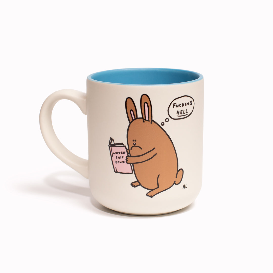 'Watership Downer' stoneware mug by Al Murphy for USTUDIO shosw a sad faced bunny rabbit thinking an expletive, whilst reading a copy of the classic animal themed novel 'Watership Down' by Richard Adams.