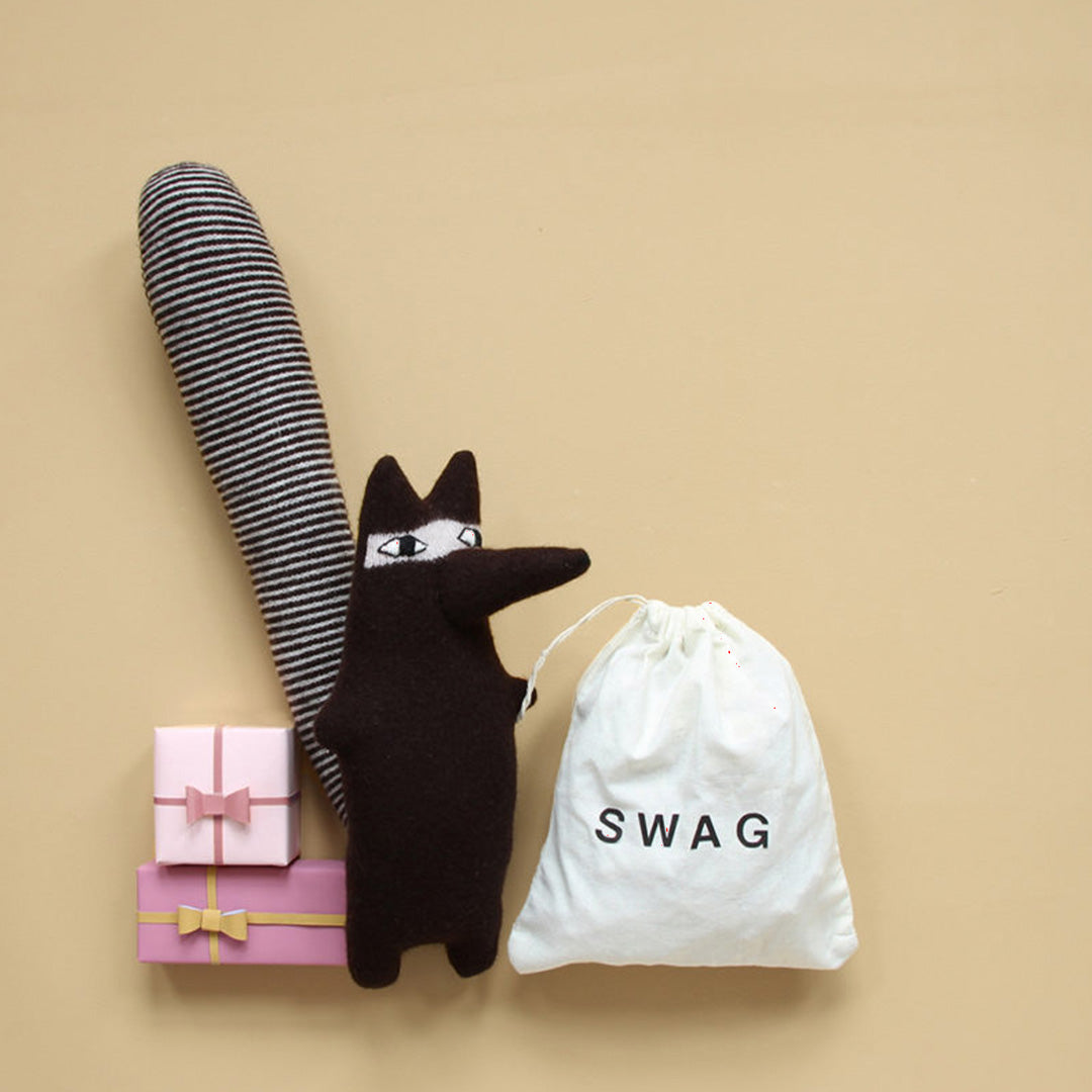 Rudie Racoon is a super-soft, sneaky thief (always on the hunt for swag), from Donna Wilson's brilliant creatures collection. Dark brown lambswool knitted raccoon in balaclava styling. lifestyle image with swag and boxes